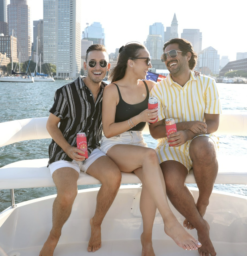Three smiling passengers enjoying drinks with their coozies for cruising available as add-ons for boat cruises in Boston